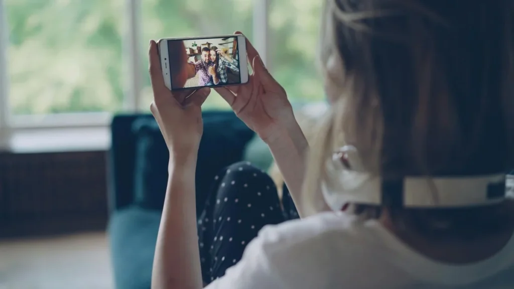 Woman sitting on couch, capturing selfie with phone indoors, wearing headphones.