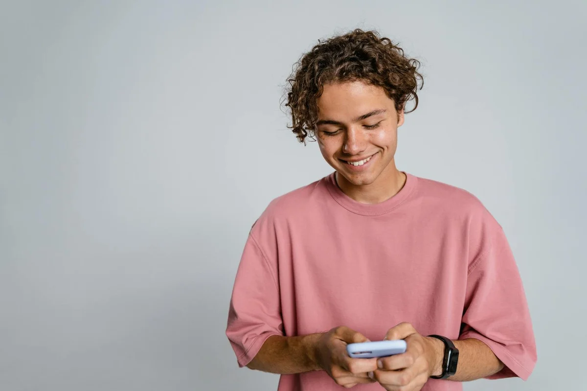 Teenager with curly hair smiling down at smartphone in a studio setup.