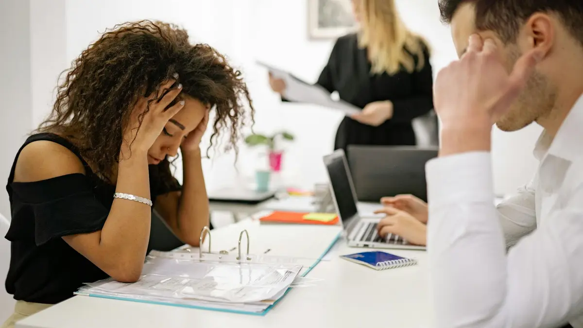African American woman stressed at office desk with colleagues nearby, conveying workplace pressure.