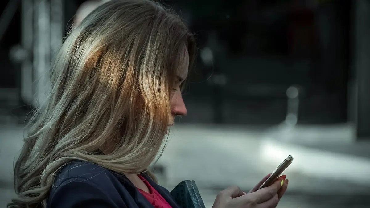 Side profile of a young woman texting on her smartphone in a shaded outdoor area.