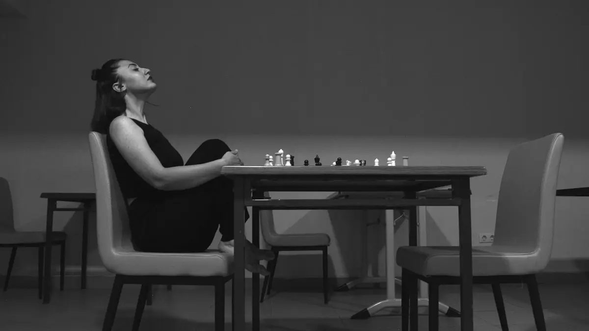A woman sits thoughtfully at a chess table in grayscale setting, evoking strategic contemplation.