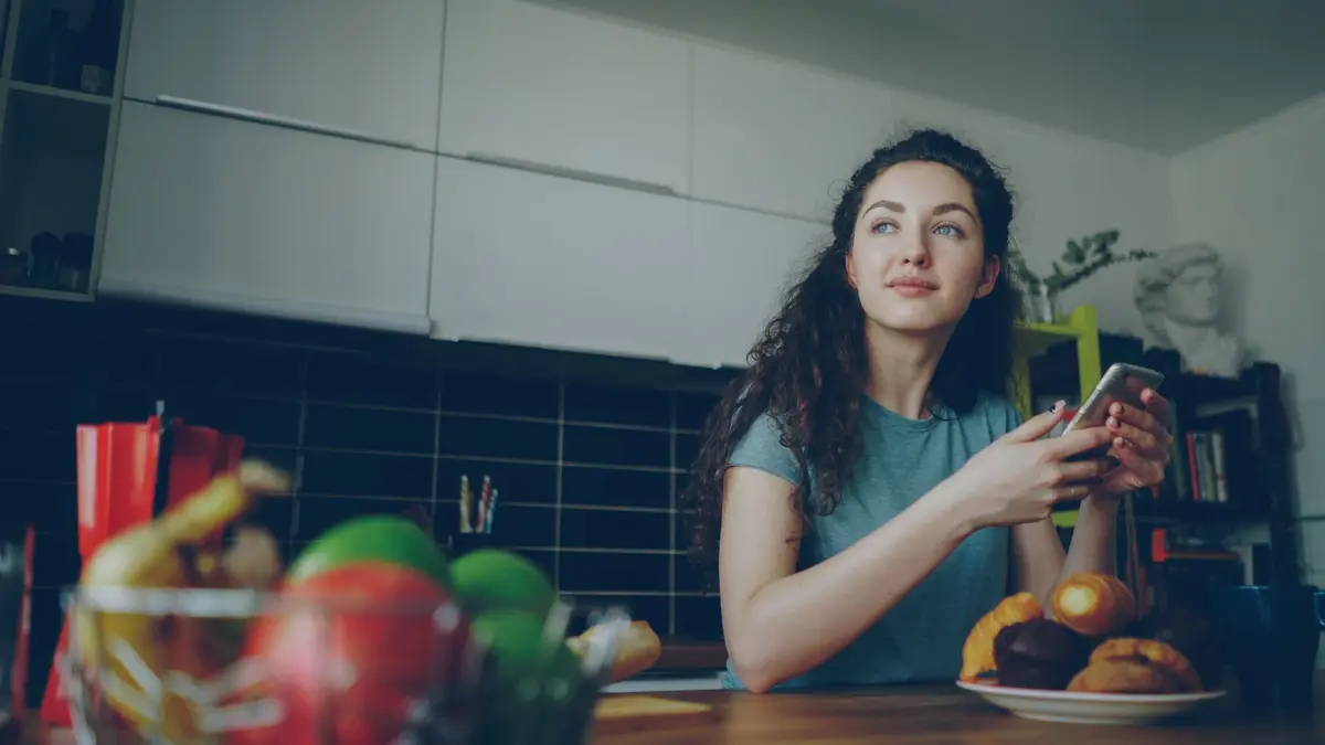 A young woman peacefully holding her smartphone while enjoying breakfast in a modern kitchen.