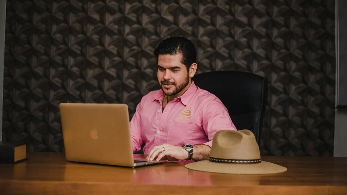 Businessman working on a laptop in an office setting, wearing a pink shirt, with a hat on the desk.