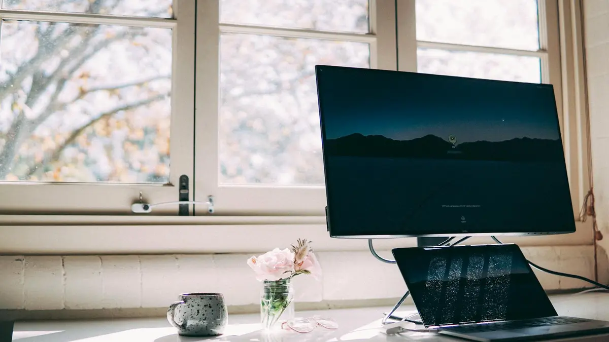 A peaceful home office setup with a desktop and laptop by a window, decorated with flowers and a mug
