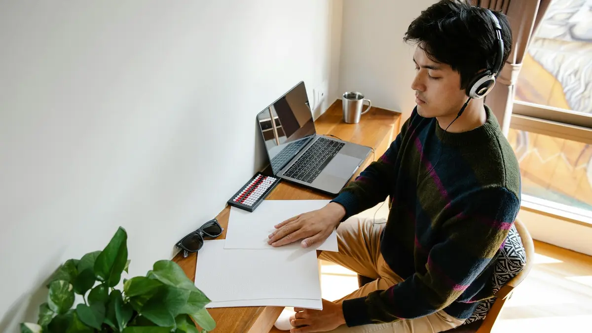 A man wearing headphones concentrates while working at a desk with plants and a laptop.